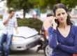 A woman stands in front of two damaged cars, talking on a phone. A man in the background leans against one of the cars, also on the phone.