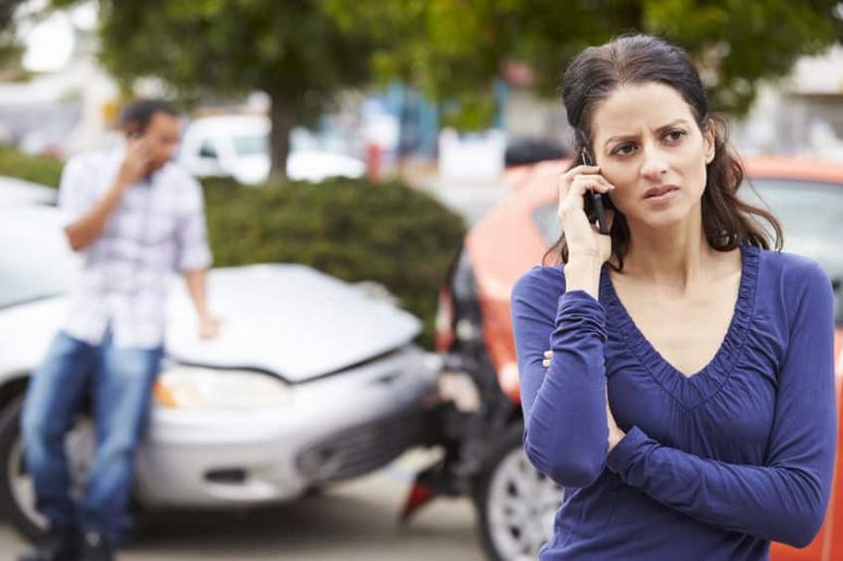 A woman stands in front of two damaged cars, talking on a phone. A man in the background leans against one of the cars, also on the phone.
