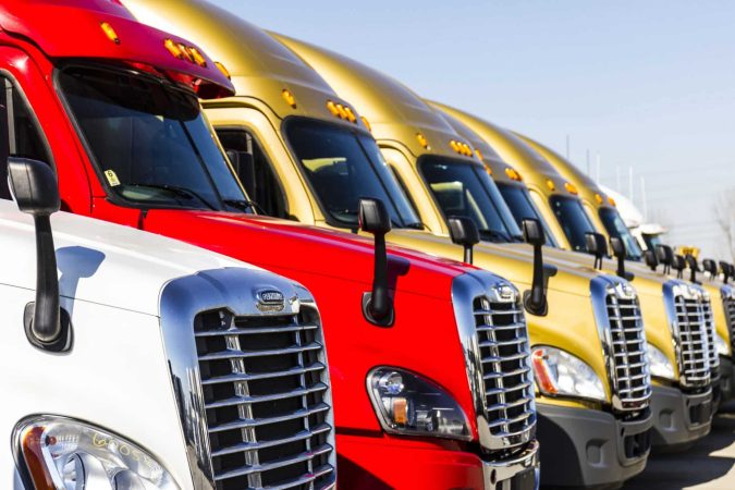 Several colorful semi-trucks, primarily red and gold, are parked in a neat row, showcasing their front grilles and headlights under a clear blue sky—perfect for anyone needing to file a truck accident claim or seeking accident information without hassle.