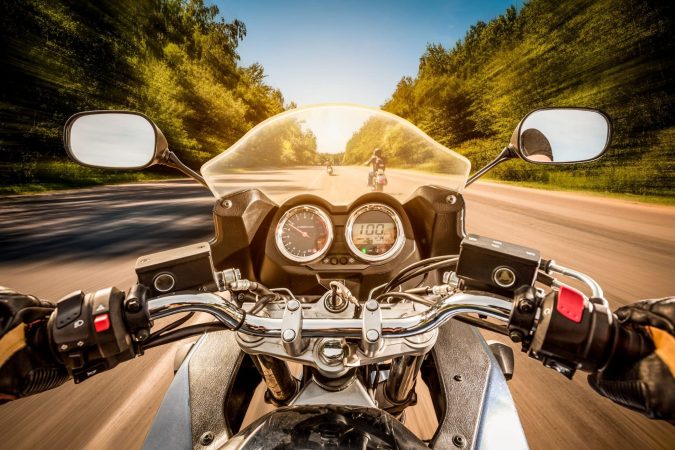 Motorcyclist's view of riding on a clear Michigan road bordered by trees, with the speedometer showing 100 km/h and another motorcyclist visible ahead.