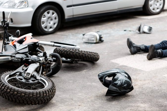 A motorcycle accident scene unfolds with a fallen bike, scattered helmet, and a person lying on the ground near a parked car. Even the safest motorcyclists face unpredictable challenges.