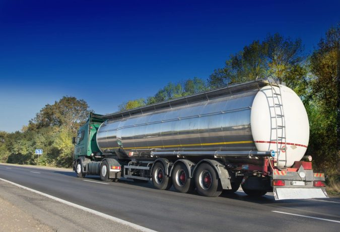 A large silver tanker truck transports through Michigan on a highway bordered by trees under a clear blue sky.