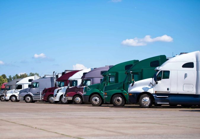 A row of colorful semi-trucks, often involved in commercial vehicle accidents, is parked side by side on a paved lot under a blue sky with scattered clouds.