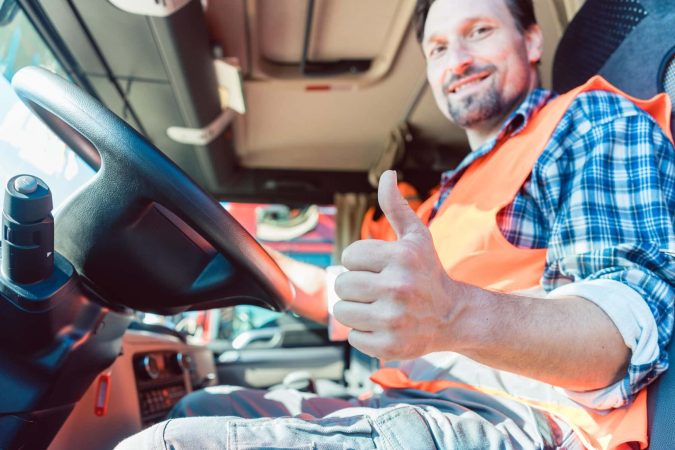 A truck driver in an orange vest gives a thumbs up from the cab, showcasing the commitment truck drivers have as they save lives daily through responsible practices like regular drug and alcohol testing.