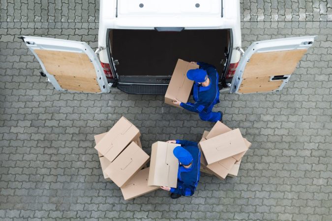 Two workers in blue uniforms carefully unload cardboard boxes from the back of a white van onto a paved surface, ensuring there is no improper loading that could lead to accidents.