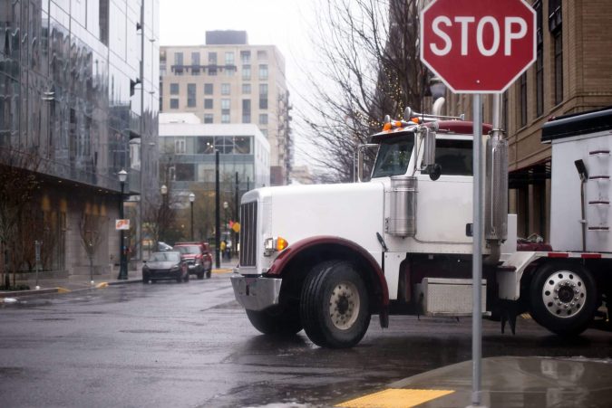 A white semi-truck makes a turn at an intersection on a rainy day in the city, navigating carefully near a stop sign. Despite the weather's dangerous conditions for pedestrians, the truck glides smoothly without incident.