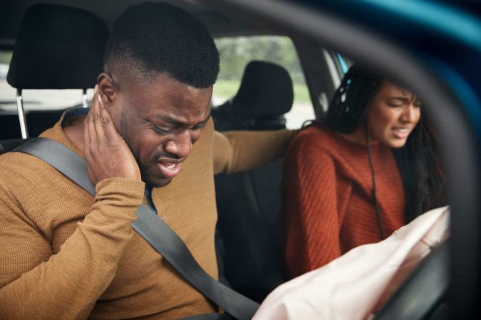 Two people sit in a car, appearing in pain after a car accident. The driver holds his neck, and the passenger looks distressed amid common injuries. An airbag is deployed.