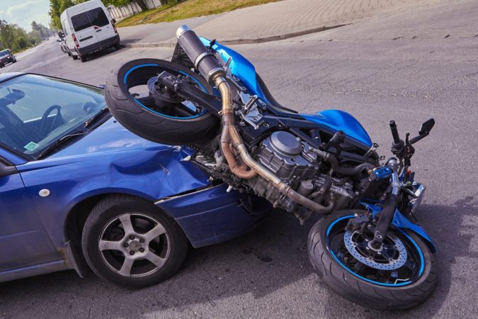 A blue motorcycle is lodged on the hood of a blue car after a collision on a Michigan street, highlighting the frequent motorcycle accidents in the area.