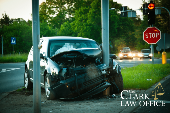 A car crashed head-on into a streetlight pole at an intersection, with front-end damage visible. The Clark Law Office logo is in the image corner.