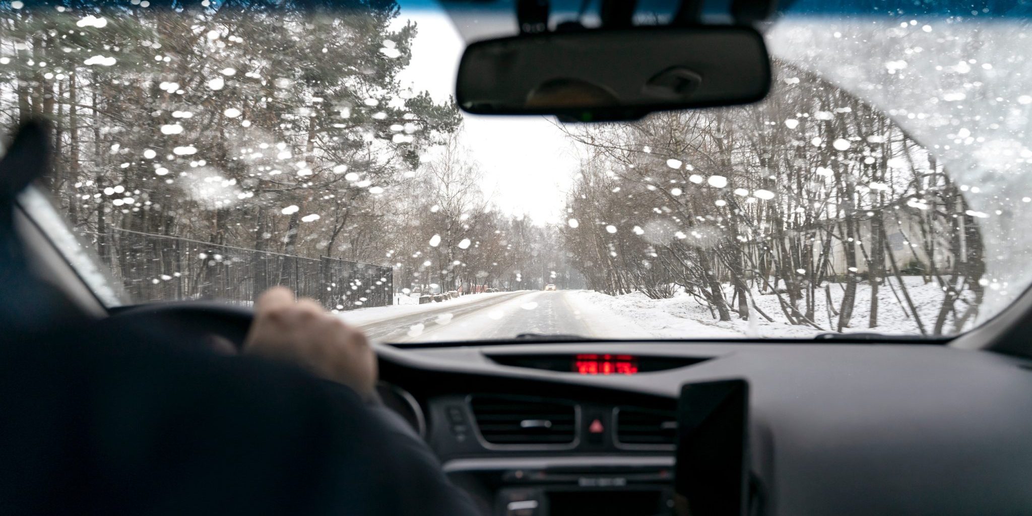 How Lansing Weather Increases The Risk of Car Accidents View from inside a car driving on a snowy road with trees on both sides. The windshield has water droplets, a reminder of the Lansing weather's potential impacts. The dashboard and steering wheel remain visible as caution drives awareness of car accident risks.