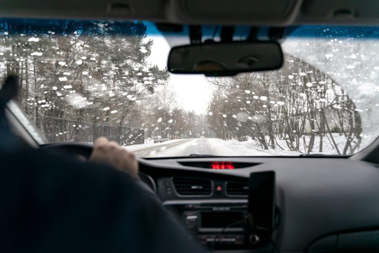 View from inside a car driving on a snowy road with trees on both sides. The windshield has water droplets, a reminder of the Lansing weather's potential impacts. The dashboard and steering wheel remain visible as caution drives awareness of car accident risks.