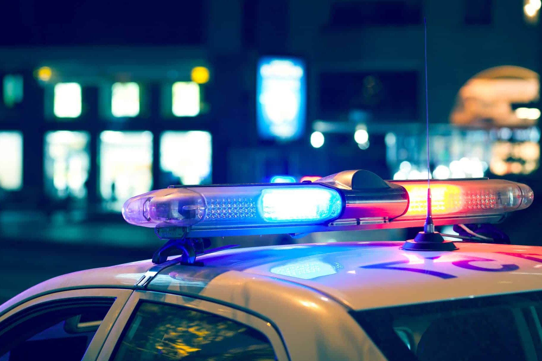 With red and blue lights flashing in the Lansing night, the police car stood parked on a city street, its presence hinting at a car accident nearby, with blurred buildings providing a mysterious backdrop.