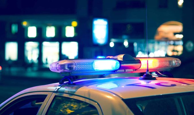 With red and blue lights flashing in the Lansing night, the police car stood parked on a city street, its presence hinting at a car accident nearby, with blurred buildings providing a mysterious backdrop.