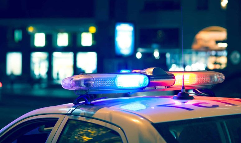 With red and blue lights flashing in the Lansing night, the police car stood parked on a city street, its presence hinting at a car accident nearby, with blurred buildings providing a mysterious backdrop.