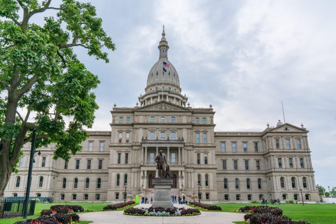 The image depicts the Michigan State Capitol building with its prominent dome, under cloudy skies. A few visitors linger near a statue in the foreground, perhaps contemplating a conversation with a local lawyer about recent events in Lansing.