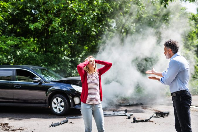 Two people stand near a damaged black car with smoke in the background on a Lansing street. A woman holds her head, showing signs of pain and suffering, while a man gestures as they discuss the car accident. Debris is scattered on the ground.