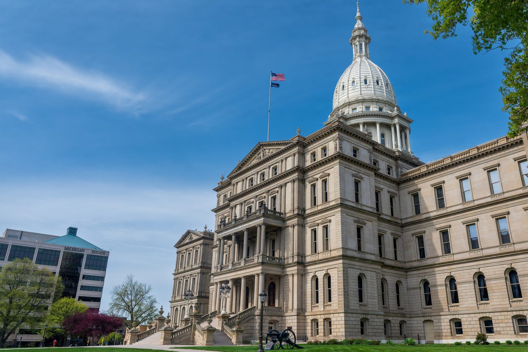 The image showcases a large historic building with a domed roof, embraced by a manicured lawn under a clear blue sky. An American flag flies proudly at the top—iconic for Lansing residents navigating their city's vibrant history like a well-versed guide.