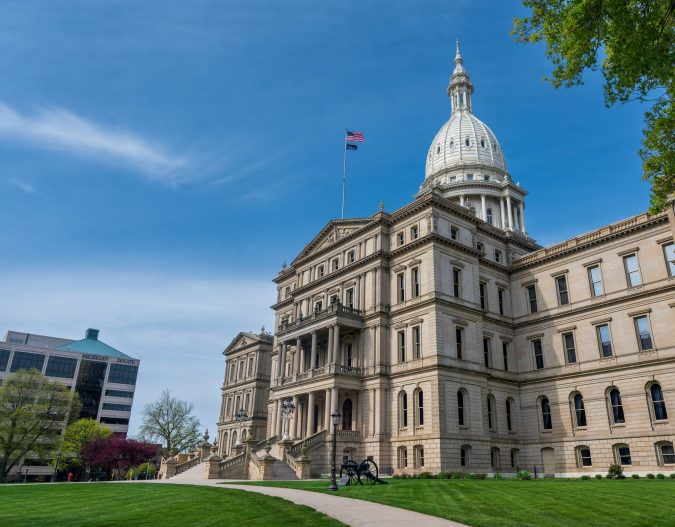 The image showcases a large historic building with a domed roof, embraced by a manicured lawn under a clear blue sky. An American flag flies proudly at the top—iconic for Lansing residents navigating their city's vibrant history like a well-versed guide.