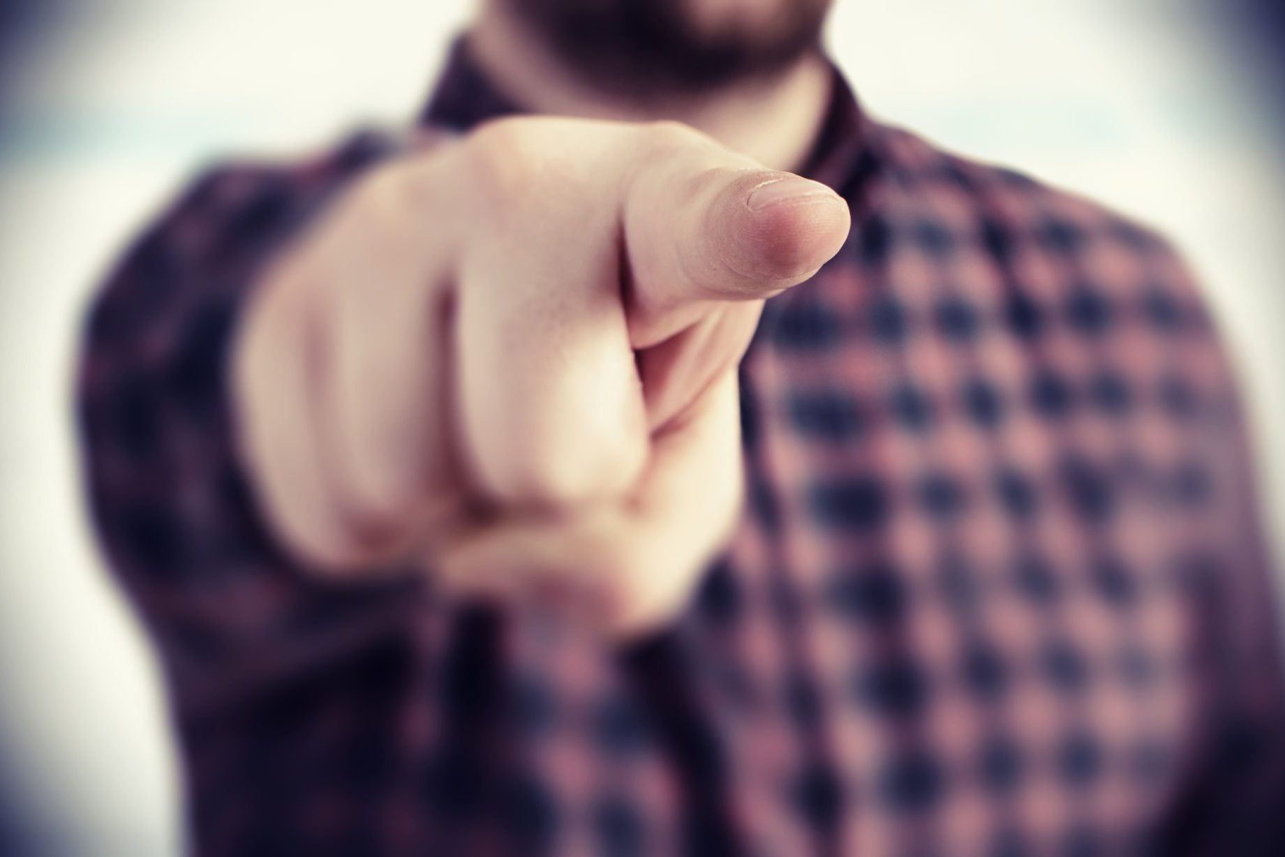 A person in a checkered shirt points their finger directly at the viewer, as if making a claim, with the background artfully blurred.