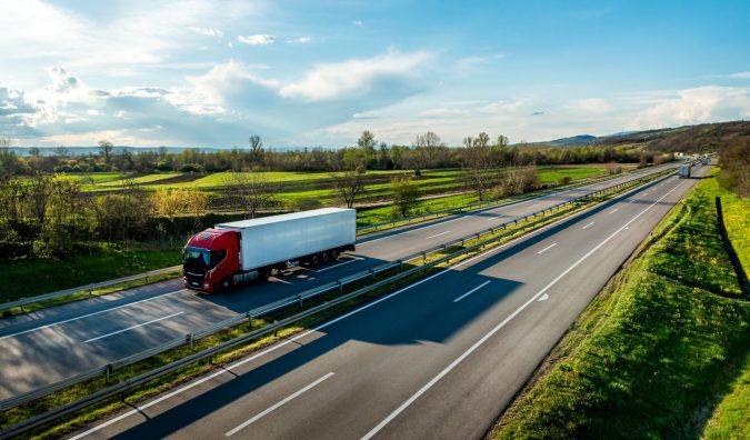 A red semi-truck with a white trailer cruises along the highway through a lush rural landscape under a partly cloudy sky, heading towards Lansing where transport professionals get paid handsomely for navigating these picturesque routes.