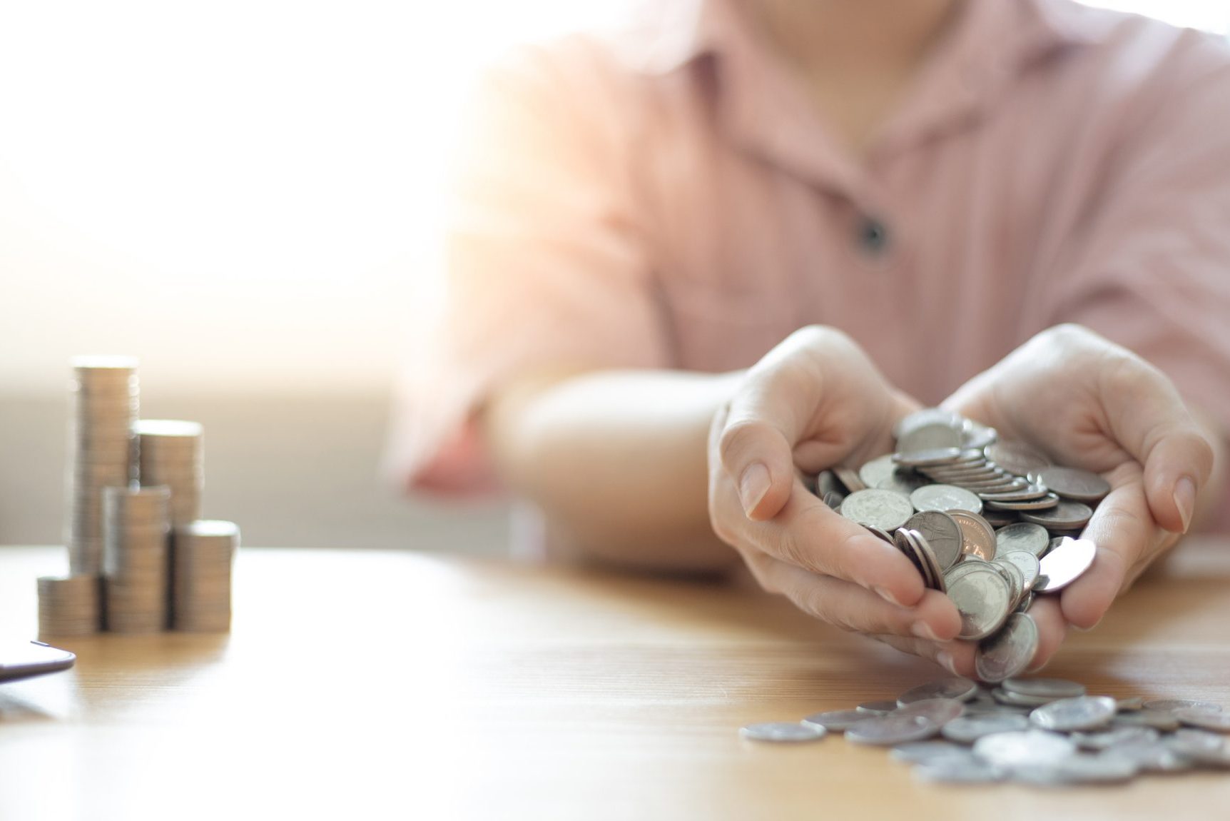 A person holding and spilling silver coins onto a wooden table, with stacks of coins in the background, as if reclaiming treasure after a battle to fight back against adversity.