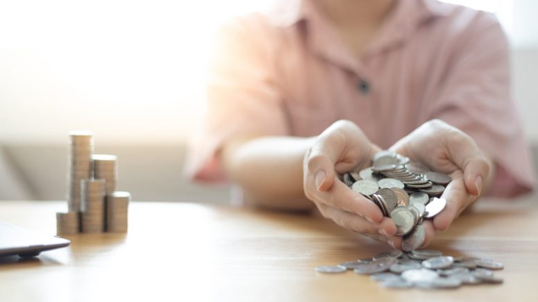 A person holding and spilling silver coins onto a wooden table, with stacks of coins in the background, as if reclaiming treasure after a battle to fight back against adversity.