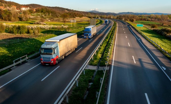 Two trucks journey on a divided highway near Lansing, flanked by green fields and rolling hills under a clear sky.