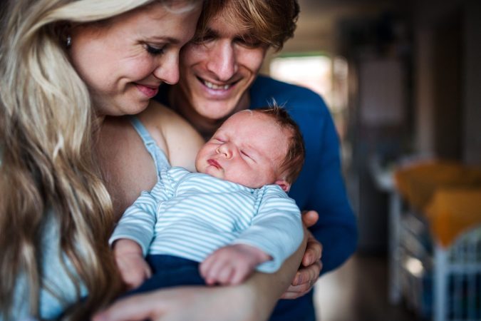 A couple in Lansing gently cradles a sleeping baby. The woman with long blonde hair and the man with short light brown hair look content and close, unaware their journey might inspire a birth injury legal guide someday. The baby, peacefully dressed in a striped shirt, completes the serene scene.