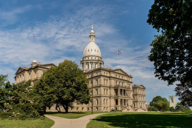 Historic building with a dome and flag in Lansing, surrounded by trees and a pathway. Bright daylight with blue sky and clouds creates a picturesque scene, reminiscent of the city's rich history.