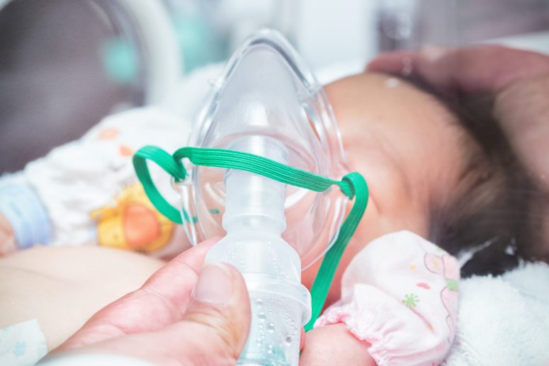Long Term Effect of Birth Asphyxia A baby, a victim of birth asphyxia, lies down with an oxygen mask on their face. A hand—perhaps one of the parents—gently holds the mask in place, concerned about potential long-term effects.