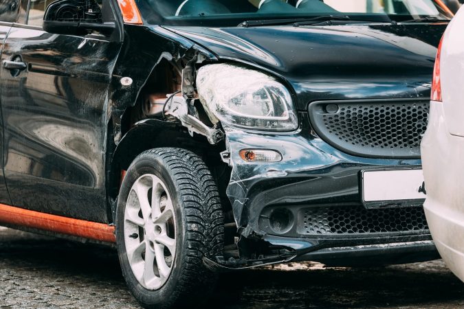 A black car with visible front-end damage, likely from a minor car accident, is parked on a Lansing street next to another vehicle.