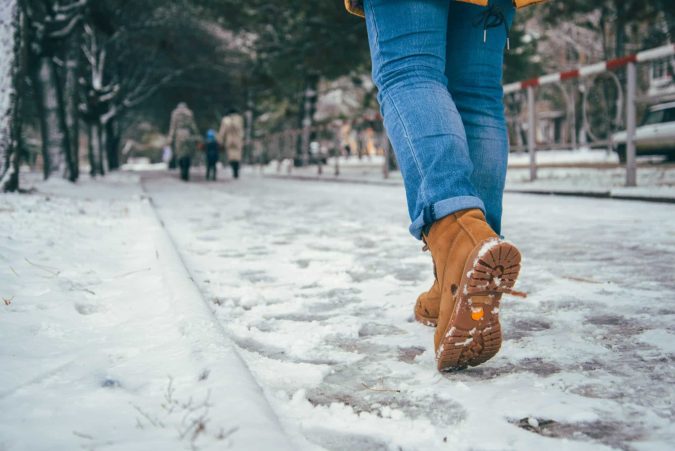 A close-up captures a person in blue jeans and tan boots cautiously navigating a snow-covered path, mindful of potential slip and fall hazards, with trees and people blurred in the background.