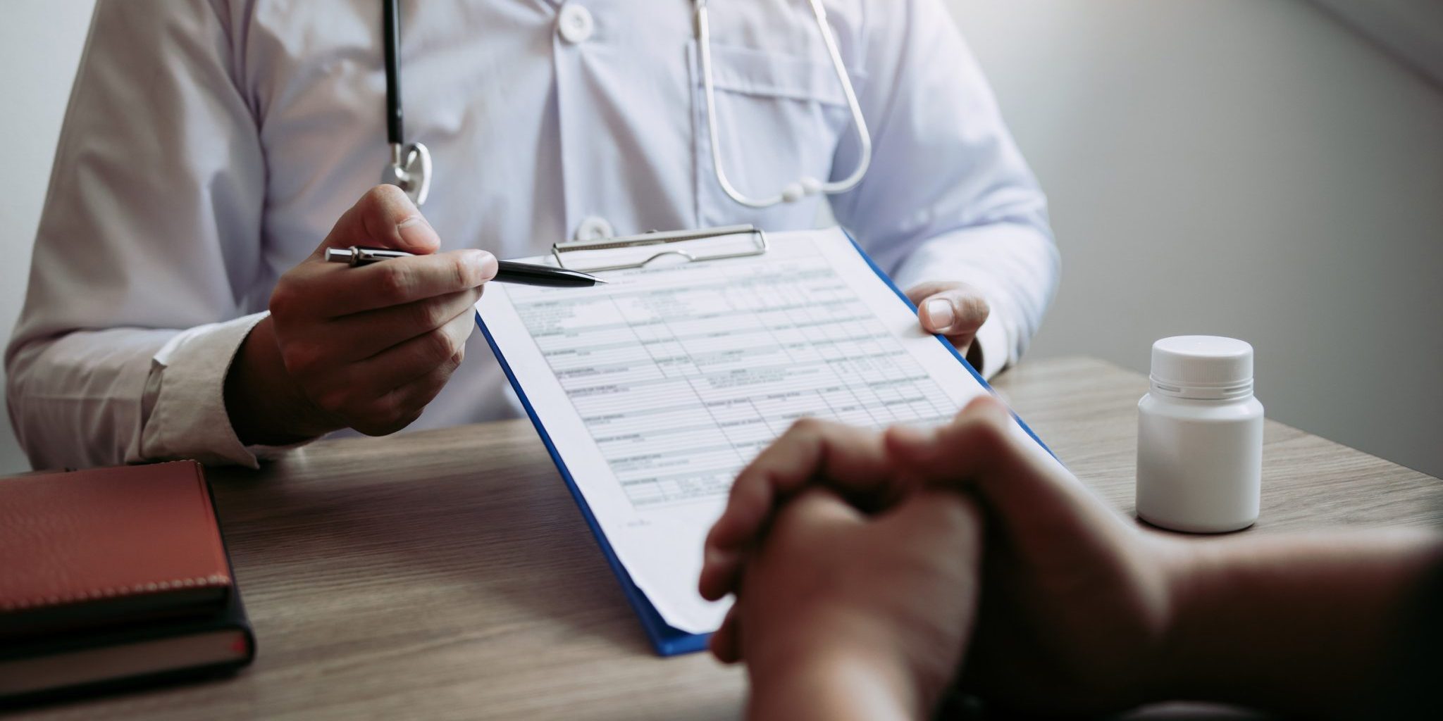 Can You Sue Urgent Care in Michigan In an urgent care setting, a doctor in a white coat holds a pen and clipboard while discussing with a patient. A bottle of pills and a notebook rest on the table, ensuring every detail is noted with precision in Michigan's trusted healthcare environment.