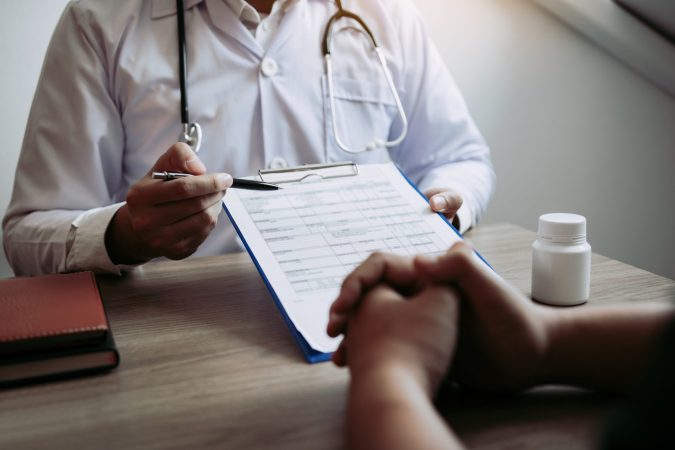 In an urgent care setting, a doctor in a white coat holds a pen and clipboard while discussing with a patient. A bottle of pills and a notebook rest on the table, ensuring every detail is noted with precision in Michigan's trusted healthcare environment.