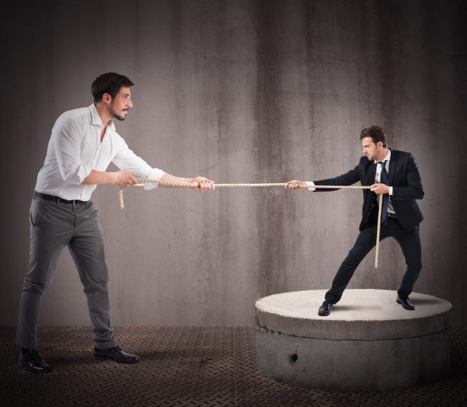 In a symbolic showdown of local vs. big firm, two men engage in a tug-of-war. The man on the left, wearing a white shirt, stands firmly on the ground, while his suited rival stands on a raised circular platform.