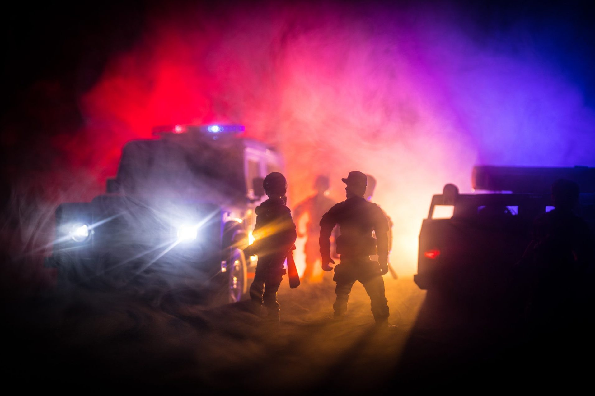Silhouettes of toy action figures stand defiantly in front of toy police vehicles with headlights cutting through the red and blue mist, evoking the chaotic aftermath of car accidents.