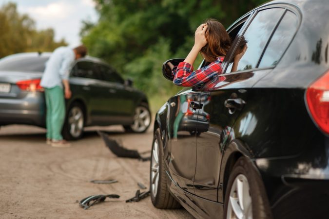 Two cars collided on a rural road near Lansing. A woman in a plaid shirt sits distressed in one car, while a man examines the damage to the other. Debris from the car accident is scattered across the ground, highlighting the cost of unexpected mistakes.