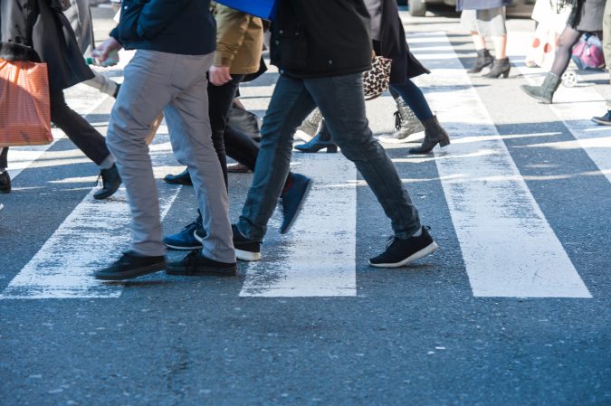 People walking across a crosswalk on a city street in Lansing, visible from the knees down, with various shoes and clothing styles—a common scene near MSU and a reminder to stay alert for pedestrian accidents.