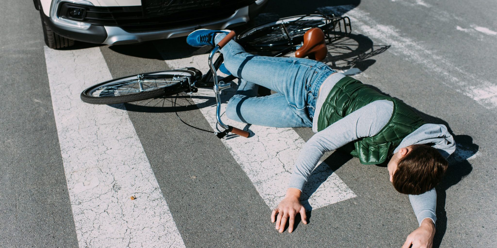 MSU Bicycle Crash A person lies on a crosswalk next to a bicycle, partially under the front of a car, appearing to have suffered a biking injury in what looks like an MSU crash.
