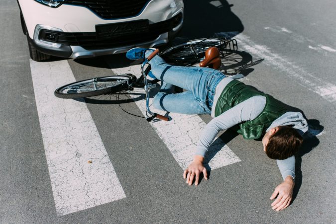 A person lies on a crosswalk next to a bicycle, partially under the front of a car, appearing to have suffered a biking injury in what looks like an MSU crash.