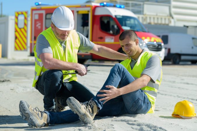 Two construction workers in safety vests and helmets; one is sitting and holding his injured leg while the other assists him, highlighting a hurt on the job situation in Lansing with an ambulance in the background.