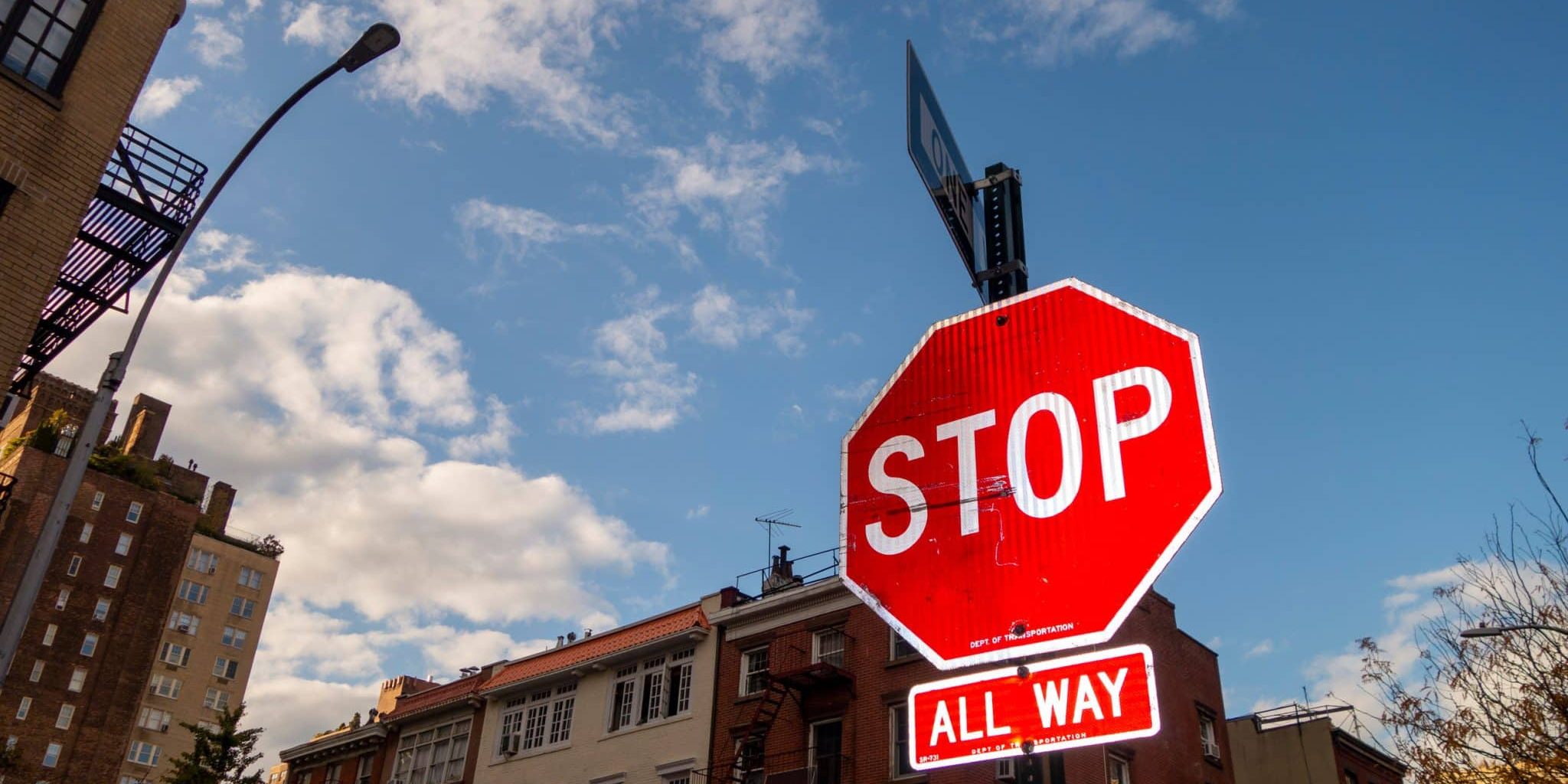 A red stop sign with an “All Way” plaque below it stands at a street corner in Michigan, highlighting a typical 4-way stop where Michigan traffic laws determine who is at fault at the intersection. Buildings and a partly cloudy sky are seen in the background.