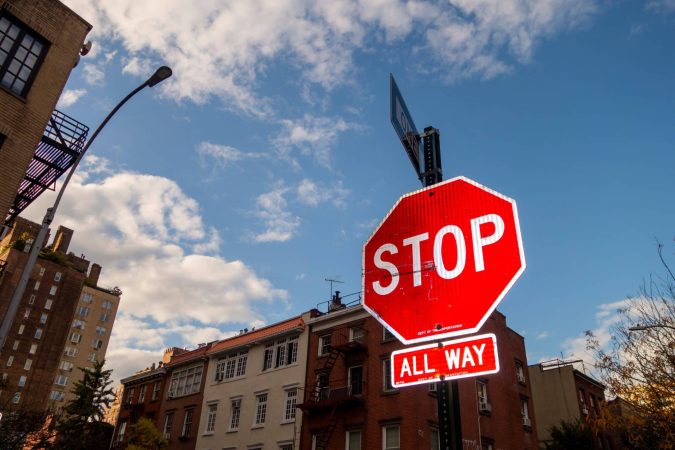 A red stop sign with an “All Way” plaque below it stands at a street corner in Michigan, highlighting a typical 4-way stop where Michigan traffic laws determine who is at fault at the intersection. Buildings and a partly cloudy sky are seen in the background.