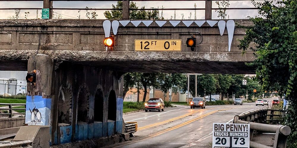 big-penny-bridge-lansing The Big Penny Bridge in Lansing features a 12-foot clearance sign, warning lights, painted teeth decorations, and a sign tallying truck wrecks. Cars drive below this unique concrete railway bridge.