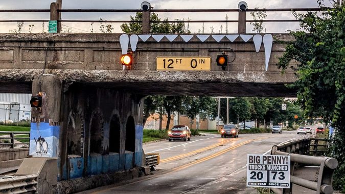 The Big Penny Bridge in Lansing features a 12-foot clearance sign, warning lights, painted teeth decorations, and a sign tallying truck wrecks. Cars drive below this unique concrete railway bridge.