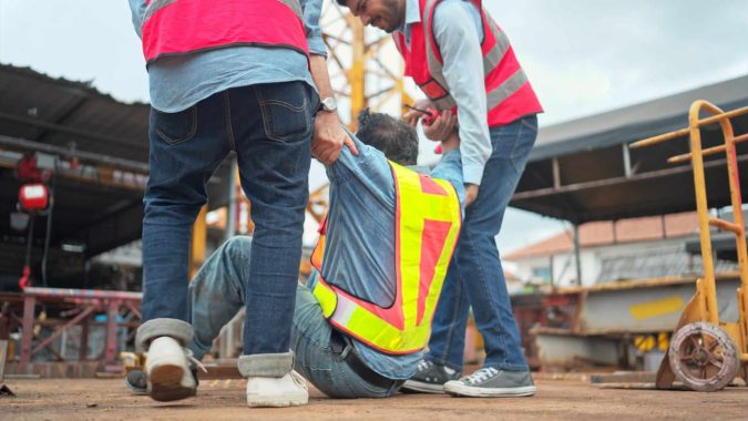 Two workers in safety vests assist a colleague who has fallen on a construction site, offering support that may be crucial in a Workers' Comp Lansing claim.