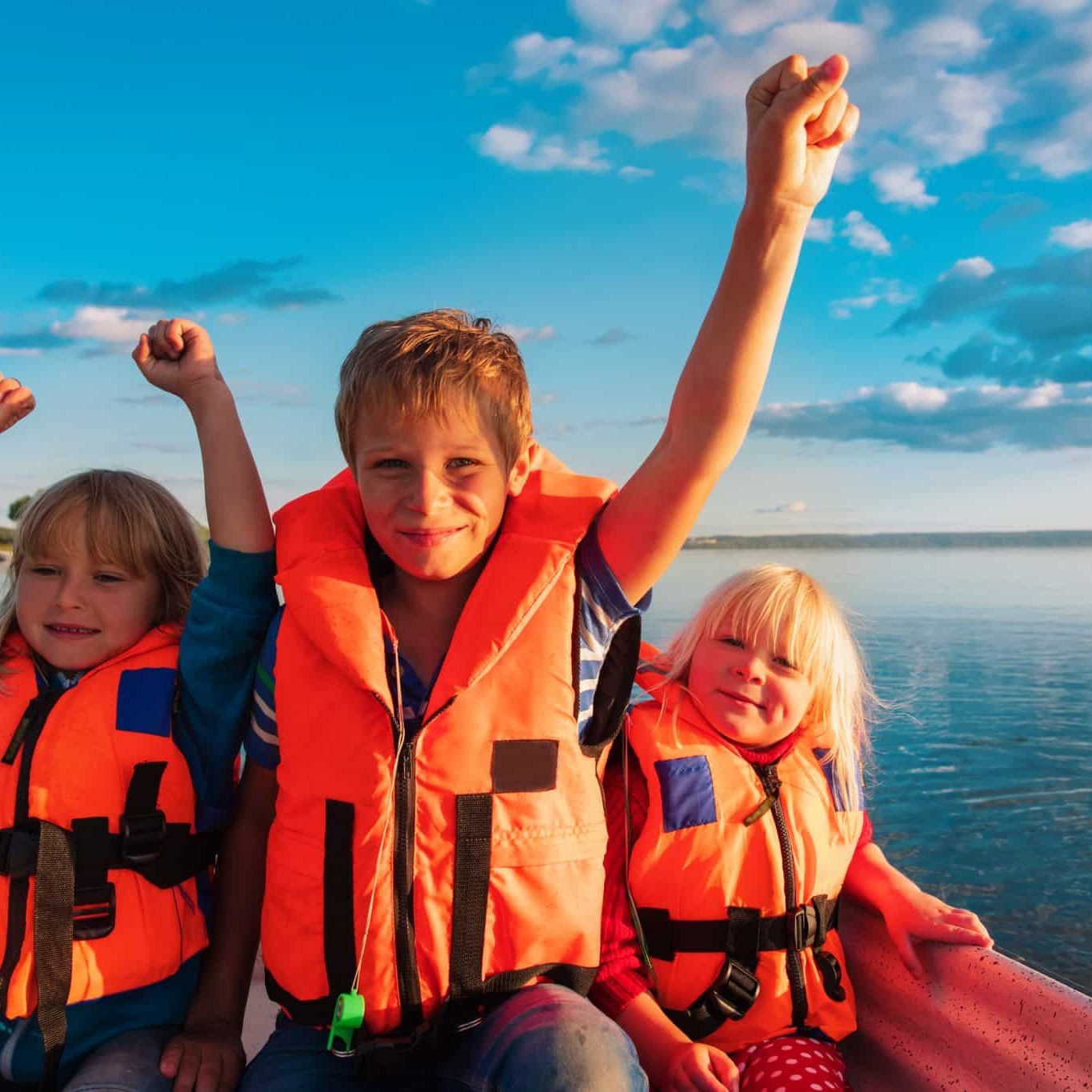 Criminal Homicide vs. Civil Wrongful Death in Michigan Three children wearing orange life jackets, following Michigan life jacket laws, sit in a boat on a lake, two with arms raised, under a blue sky with scattered clouds.
