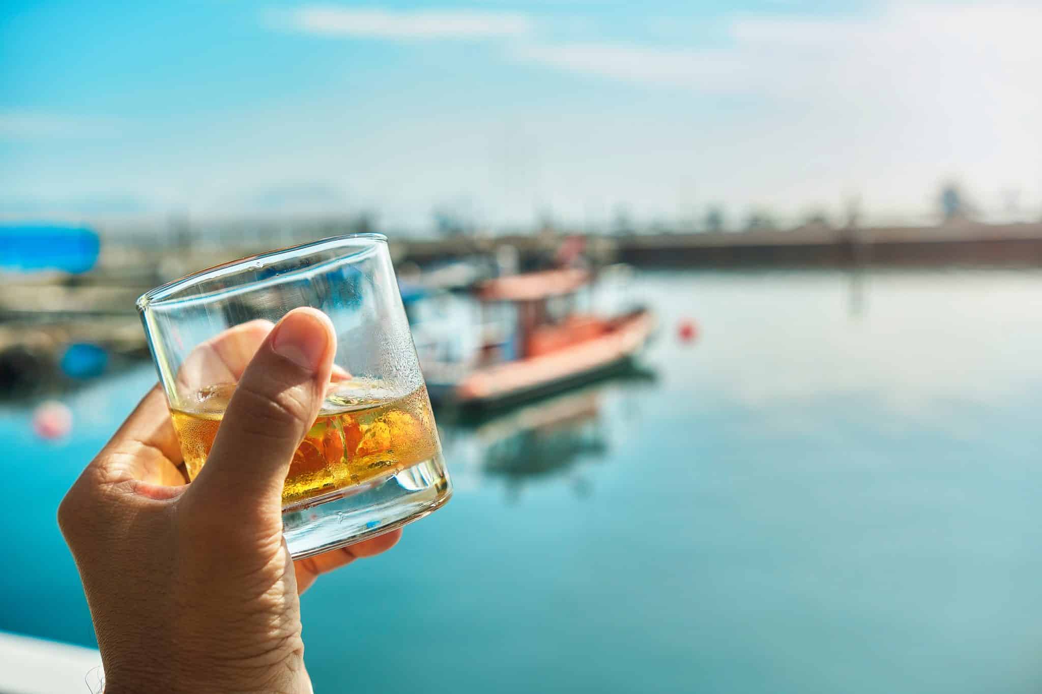 A hand holding a glass of whiskey with ice, with a blurred view of boats and water in the background—reminding us to follow safe boating Michigan guidelines while enjoying the view.