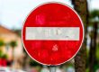 A red and white "Do Not Enter" traffic sign, symbolizing common workers’ comp mistakes, is faded and covered with worn stickers, positioned outdoors with trees and buildings in the blurred background.