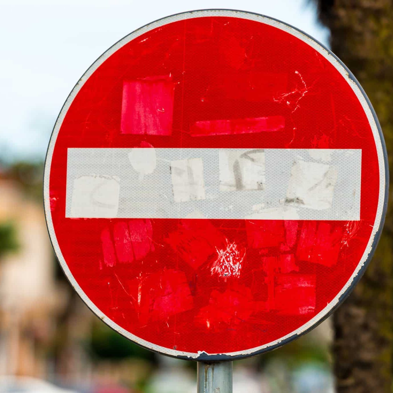 A red and white "Do Not Enter" traffic sign, symbolizing common workers’ comp mistakes, is faded and covered with worn stickers, positioned outdoors with trees and buildings in the blurred background.